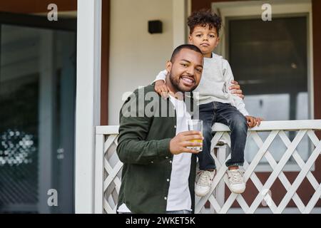 gioioso padre afroamericano in bretelle che tengono il succo d'arancia e abbracciano suo figlio seduto sul portico Foto Stock