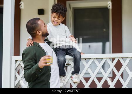 allegro padre afroamericano in bretelle che tengono il succo d'arancia e abbracciano suo figlio seduto sul portico Foto Stock