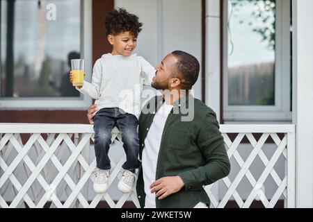 gioioso uomo afroamericano in bretelle che tiene il succo d'arancia e abbraccia suo figlio seduto sul portico Foto Stock