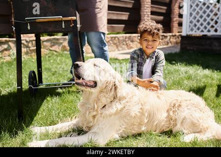 sorridente ragazzo afro-americano seduto vicino al cane mentre il padre cucina barbecue sul cortile di campagna in periferia Foto Stock