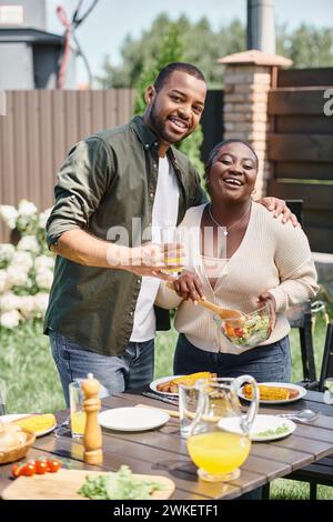 felice uomo afro-americano che abbraccia moglie mescolando insalata in una ciotola di vetro mentre si gusta un barbecue sul cortile Foto Stock