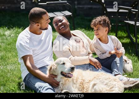 momenti di famiglia con genitori afroamericani e figli sorridenti e seduti sul prato verde vicino al cane Foto Stock