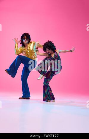 Ballerini entusiasti, uomo e donna in un colorato abito anni '70 che danzano in movimento su uno sfondo rosa sfumato in studio. Foto Stock