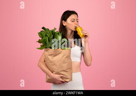 Indossa un abito sportivo bianco che sorseggia succo d'arancia e tiene in mano un sacchetto di carta pieno di verdure verdi Foto Stock