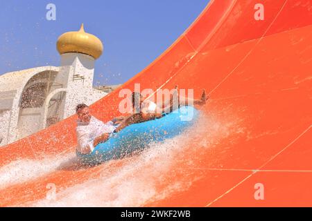 Famiglia che scende su uno scivolo d'acqua in un parco acquatico in estate. Spruzzi d'acqua. Padre e figlia piccola cavalcano su anelli gonfiabili in vacanza al S Foto Stock