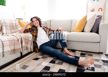 Foto di un ragazzo allegro di buon umore vestito camicia a scacchi che si siede su un tappeto che si gode il fine settimana in casa Foto Stock