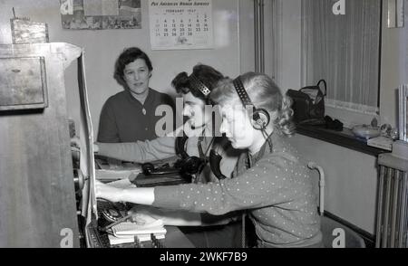 1957, storico, all'interno di una stanza, due ragazze, che indossano le cuffie, imparare a gestire un centralino telefonico, essere supervisionato da una signora seduta accanto a loro. Sul muro, un calendario della Pan American World Airways, datato febbraio 1957. Su uno scaffale ci sono direzioni telefoniche per le regioni di Outer London, West Middlesex, Middlesex e Kent. Foto Stock