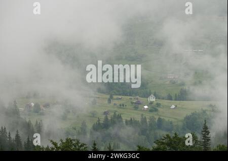 Fitta nebbia tra le montagne, attraverso la quale case e il villaggio sono appena visibili, foschia mattutina tra le montagne. Foto Stock