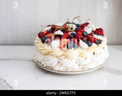 Torta Pavlova con formaggio cremoso montato, salsa al cioccolato, fragole fresche, mirtilli e lamponi, decorata con meringhe su sfondo semplice Foto Stock
