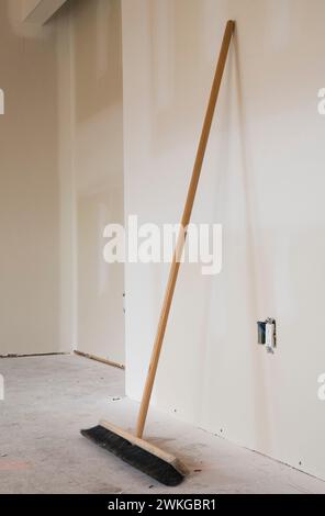 Scopa spazzolante appoggiata su pareti in cartongesso non verniciate in una stanza incompiuta con pavimento in compensato polveroso all'interno della casa. Foto Stock