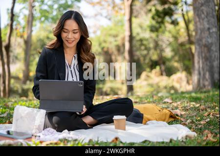 Una donna d'affari asiatica positiva e attraente che lavora a distanza in un parco, lavorando sul suo tablet digitale su un tappetino da picnic. Foto Stock