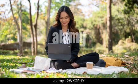 Una donna d'affari asiatica positiva e attraente che lavora a distanza in un parco, lavorando sul suo tablet digitale su un tappetino da picnic. Foto Stock