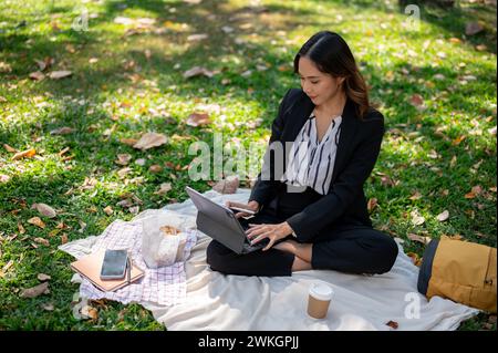 Una donna d'affari asiatica positiva e attraente che lavora a distanza in un parco, lavorando sul suo tablet digitale su un tappetino da picnic. Foto Stock