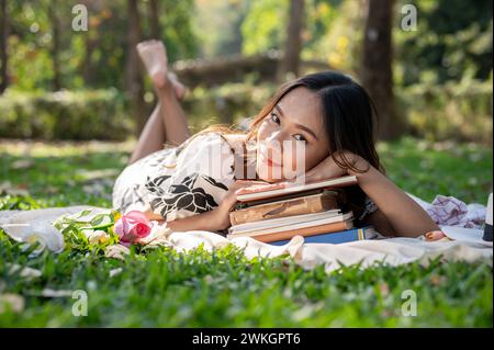 Un'attraente donna asiatica è sdraiata su un tappeto da picnic, mette la testa su una pila di libri e si gode un picnic in un giardino verde o in un parco luminoso Foto Stock