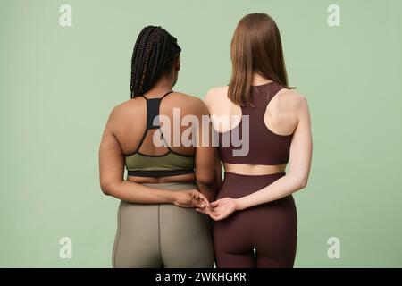 Ripresa in studio con vista posteriore di irriconoscibili donne afroamericane e caucasiche che indossano abbigliamento sportivo tenendosi per mano dietro la schiena Foto Stock