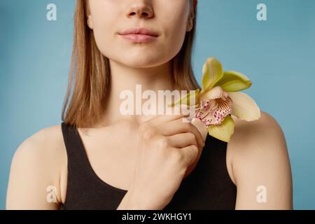 Ripresa di un raccolto medio ravvicinato di una giovane donna caucasica che tiene fresco fiore di orchidea in posa per la macchina fotografica Foto Stock