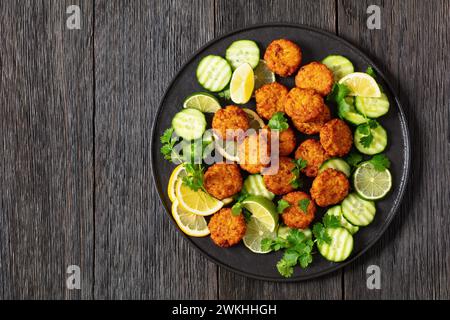 torte di riso fritte, tikki di riso, piatto indiano su piatto nero servito con cetriolo, limone, fette di lime su un tavolo di legno scuro, vista orizzontale dall'alto, f Foto Stock