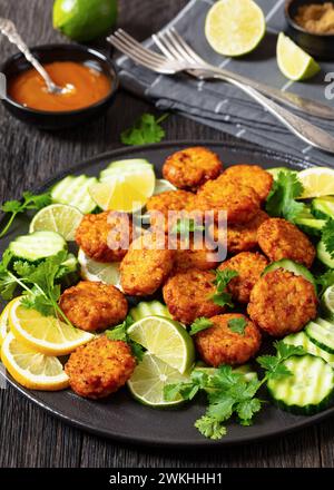 polpettine di riso fritte, tikki di riso, piatto indiano su piatto nero servito con cetriolo, limone, fette di lime e salsa al peperoncino dolce su un tavolo di legno scuro, verti Foto Stock