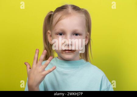 Una bambina di sette anni con una bocca sporca di cioccolato su sfondo giallo. Concetto di bambini con golosi, livelli di zucchero nel sangue, diabete e. Foto Stock
