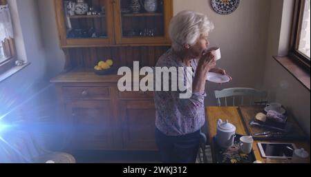 Immagine della luce che si muove sopra una donna caucasica anziana in cucina che guarda fuori dalla finestra bevendo tè Foto Stock