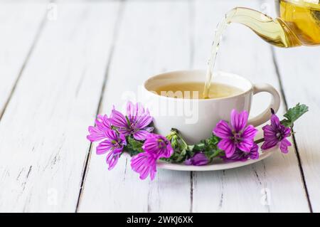 Tazza di tè al malto comune con pianta fresca di malva sylvestris in fiore su tavola rustica bianca, medicina alternativa Foto Stock