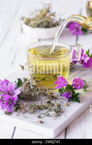 Tazza di tè al malto comune con pianta fresca di malva sylvestris in fiore su tavola rustica bianca, medicina alternativa Foto Stock