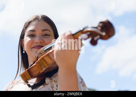 Ritratto di una giovane violinista venezuelana latina che sorride all'aperto con il suo violino che posa guardando la macchina fotografica, con il cielo sullo sfondo e copia spazio. Foto Stock