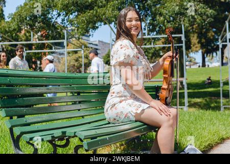 ritratto di una giovane e graziosa violinista latina che siede con il suo strumento su una panchina di legno in piazza sotto il sole, sorridente felice e guardando c Foto Stock