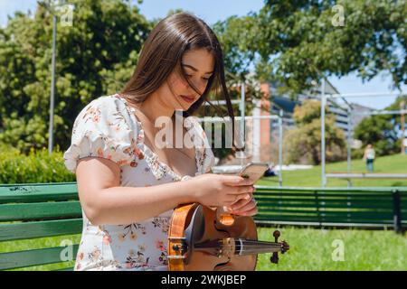 la giovane violinista latina buske seduta su una panchina di legno all'aperto, controlla tranquillamente i social network utilizzando il telefono, la connessione di persone, musica e tecnologia Foto Stock