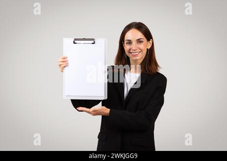 Una giovane donna professionista con una tuta nera presenta con sicurezza un portadocumenti bianco vuoto con un sorriso Foto Stock