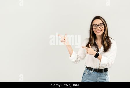 Donna felice con occhiali che puntano le dita verso un lato, indossando una camicia bianca e jeans Foto Stock