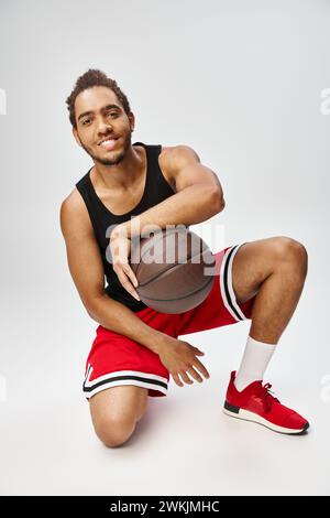gioioso e affascinante sportivo afroamericano che posa con il basket e sorride alla macchina fotografica Foto Stock