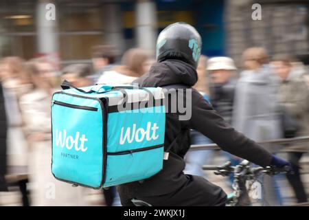 Belgrado, Serbia - 20 febbraio 2024: Servizio di corriere Wolt con una borsa di consegna in bicicletta in una strada trafficata piena di pedoni, vista posteriore Foto Stock