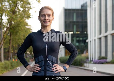 Una donna aderente all'abbigliamento sportivo con auricolari gode di un momento di riposo durante un allenamento urbano all'aperto, trasudando sicurezza e benessere. Foto Stock