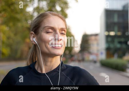 Donna sorridente con gli occhi chiusi perduta nella musica mentre utilizza auricolari bianchi, godendosi una giornata di sole in città. Foto Stock
