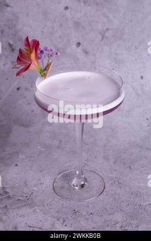 Elegante immagine di un cocktail rosa in un bicchiere con decorazione floreale su sfondo grigio, trasuda classe e raffinatezza. Foto Stock