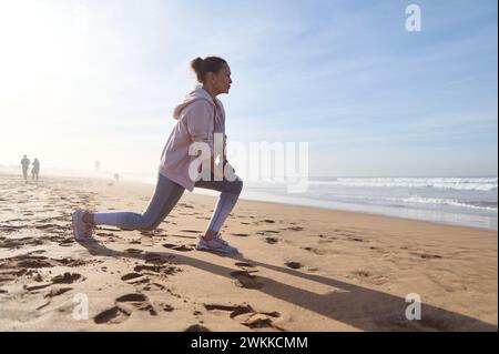 Donna sportiva attiva in abbigliamento sportivo che si esercita sulla spiaggia sabbiosa, fa affondamenti mentre riscalda il corpo, allunga i muscoli delle gambe durante l'allenamento mattutino Foto Stock