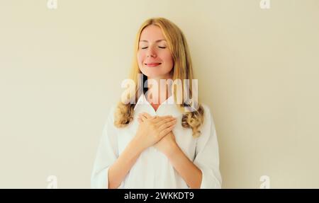 Ritratto di una donna caucasica grata bella sorridente e felice che ha messo le mani piegate sul cuore su uno sfondo bianco dello studio Foto Stock