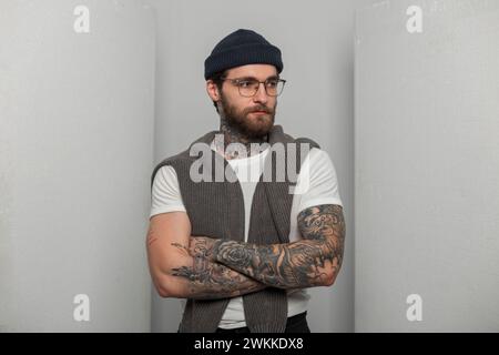 Uomo hipster dal fascino cool con barba, baffi e tatuaggi con eleganti occhiali che indossano un cappello a maglia in una t-shirt bianca alla moda con un maglione Foto Stock