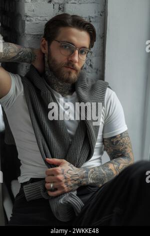 Elegante giovane hipster con tatuaggio con acconciatura e barba con occhiali alla moda in una T-shirt bianca con maglione si siede vicino alla finestra Foto Stock