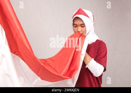 ritratto di donna asiatica hijab che bacia bandiera rossa e bianca indonesiana su sfondo bianco Foto Stock