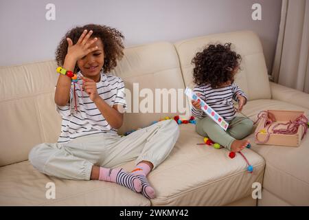 Due ragazze afroamericane in camicie a righe giocano su un divano, la più anziana con un braccialetto colorato, divertiti a imparare con giocattoli ecologici. Pol. Di alta qualità Foto Stock