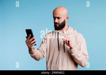Un uomo arabo serio che parla in videoconferenza su smartphone e ne mostra uno con l'indice. Persona che punta mentre ha una comunicazione remota utilizzando l'app per videocall del telefono cellulare Foto Stock