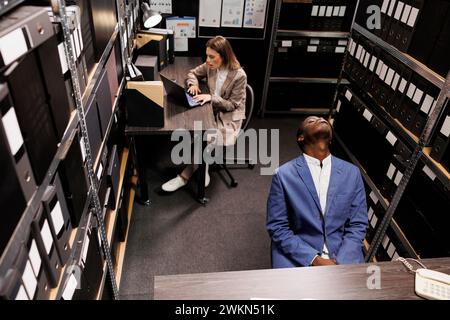 Vista dall'alto del detective privato che ha un'espressione pensiva, idee di brainstorming per risolvere il caso del crimine in sala arhive. Diversi investigatori lavorano straordinari alle indagini, analizzando nuove prove Foto Stock