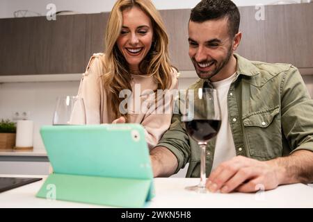 Uomo e donna felici e gioiosi in una videochiamata che beve un bicchiere di vino. Giovane coppia adulta che utilizza insieme un tablet da casa in una cucina moderna. Foto Stock