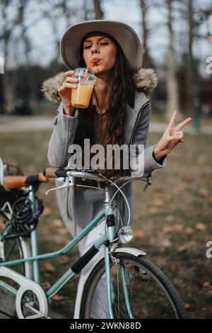 Una giovane donna alla moda sorseggia un frullato e mostra un segno di pace mentre si appoggia sulla sua bici d'epoca in un parco urbano. Foto Stock