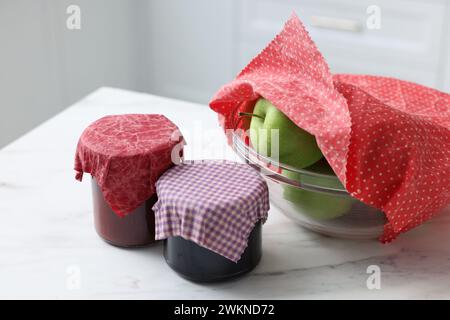 Barattoli di marmellate e mele nel recipiente ricoperti di cera d'api avvolti su un tavolo bianco all'interno, primo piano Foto Stock