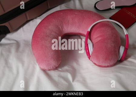 Cuscino da viaggio rosa con cuffie, valigia, passaporto e maschera dormiente sul letto Foto Stock