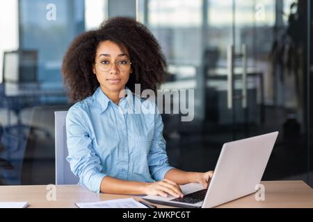 Calma signora ispanica con abiti eleganti seduti su una sedia e lavorando con un computer portatile su una scrivania in legno all'interno dell'ufficio. Segretaria attenta che prepara documenti online e prepara a completare le attività del capo. Foto Stock