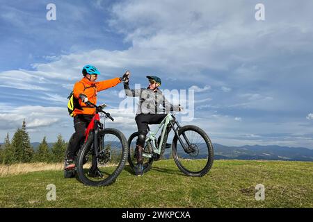 Due ciclisti uomini che guidano biciclette elettriche all'aperto. I turisti maschi che riposano sulla cima della collina, si danno cinque alti l'uno all'altro, godendo di uno splendido paesaggio montano. Concetto di svago attivo. Foto Stock